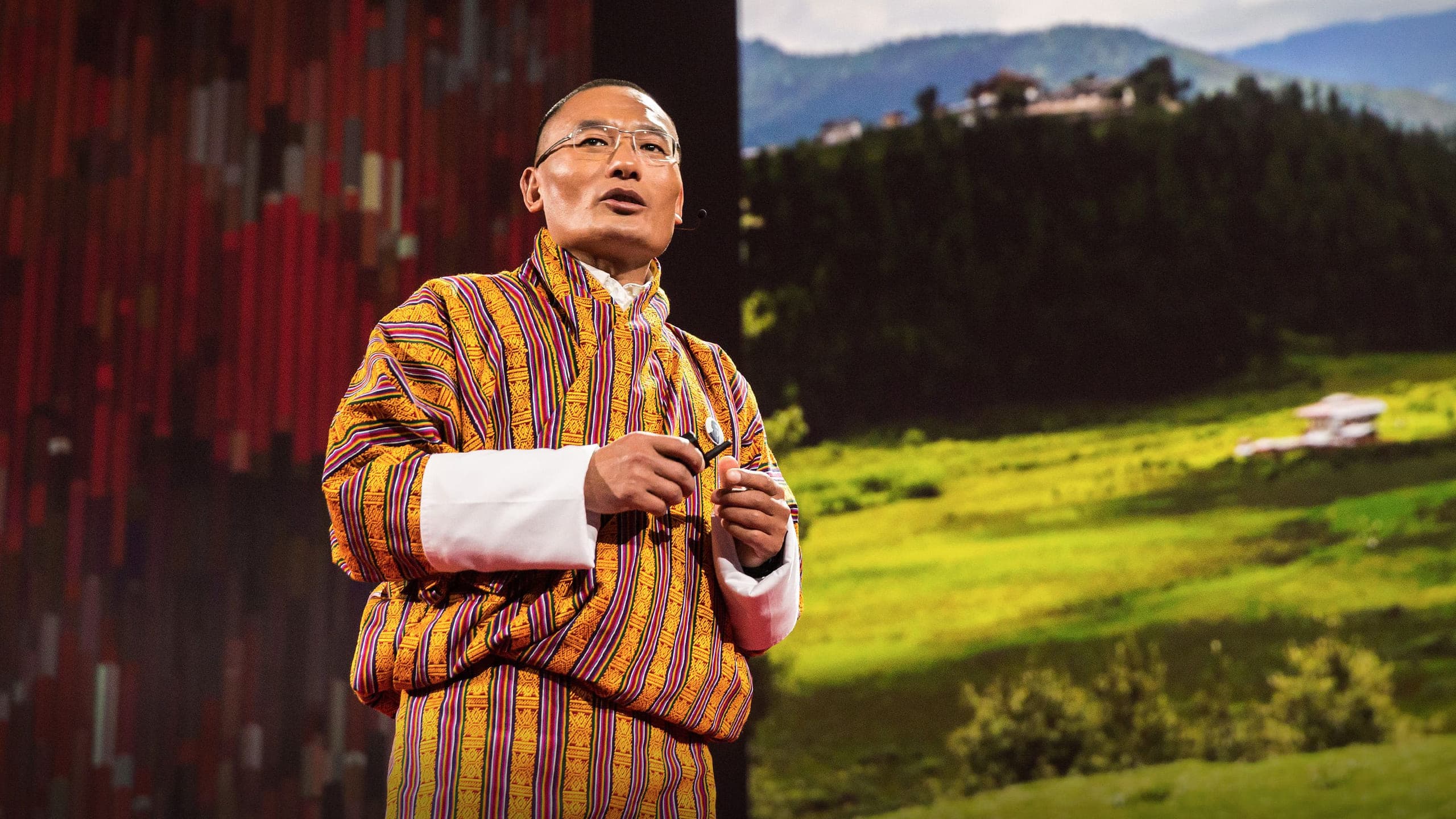 Tshering Tobgay on stage in gho, mid-talk, a Bhutanese valley projected behind him